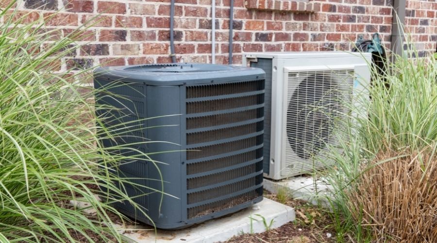 Air conditioning units on a concrete pad, surrounded by tall grasses and shrubs in a residential setting.