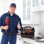 Smiling handyman with a pipe wrench and tool bag in a modern kitchen, representing Hancock & Son Plumbing services.