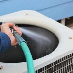 Air conditioning unit being cleaned with a garden hose, emphasizing AC coil maintenance by Hancock & Son Plumbing.