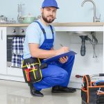 Male plumber in blue uniform working under kitchen sink, showcasing Hancock & Son Plumbing services.