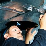 A man working on HVAC maintenance under machinery, inspecting air ducts with tools for Hancock & Son Plumbing.