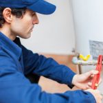 Male plumber in blue uniform adjusts plumbing valve with red wrench in utility room, representing Hancock & Son Plumbing.