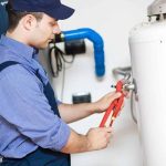 Male plumber in blue shirt fixes plumbing pipes near water heater for Hancock & Son Plumbing services.