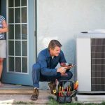 HVAC technician repairing air conditioning unit, while a relaxed observer watches from inside Hancock & Son's service area.