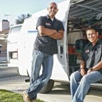 Two men in work attire next to a work van displaying tools for plumbing and HVAC services by Hancock & Son Plumbing.