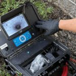 Operator inspecting plumbing inspection equipment with gloves, displaying control interface and tools in durable case.