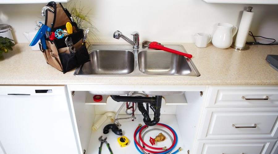 Kitchen sink with plumbing tools, including a red pipe wrench, under-sink plumbing components, and a tool bag, illustrating plumbing maintenance and repair services.